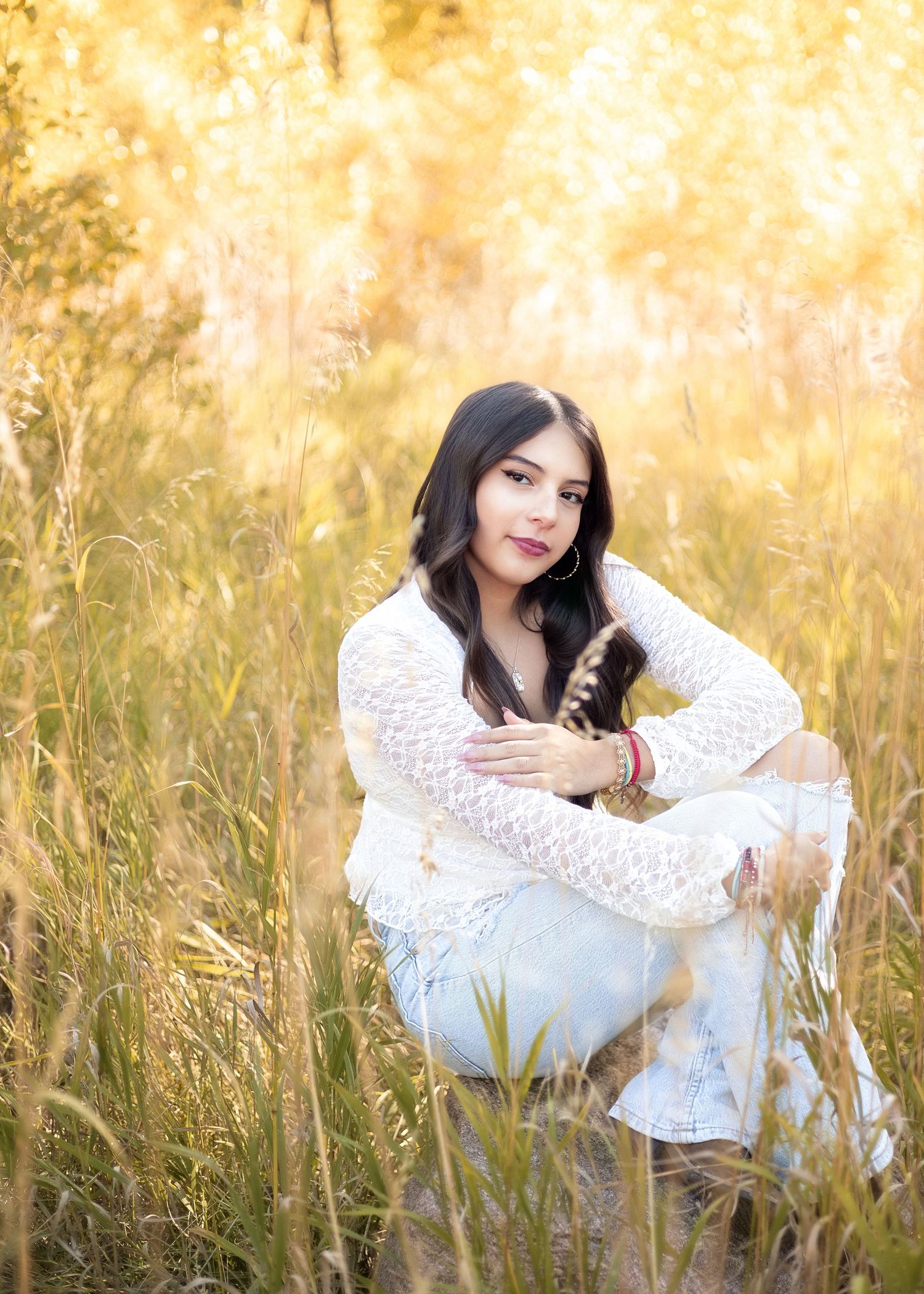 Young woman in white lace top and light jeans sitting on a log in a golden autumn meadow outdoors.