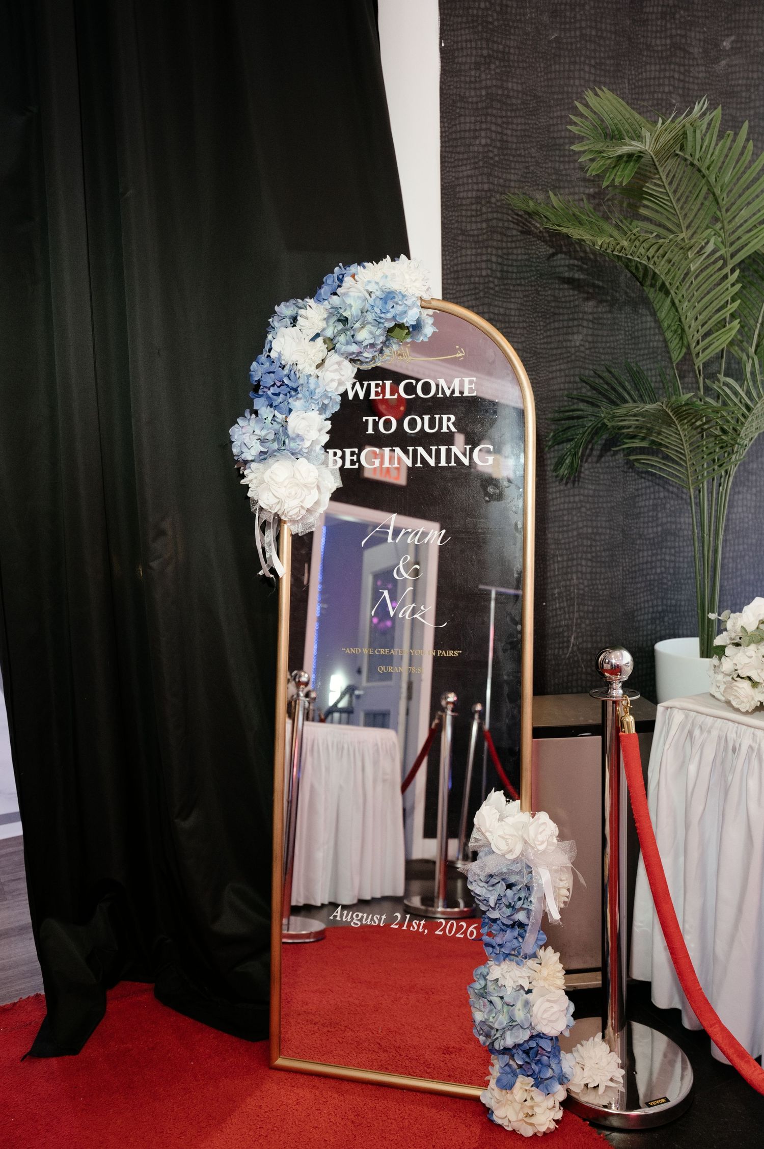 Arched gold-framed wedding welcome mirror with blue and white hydrangea florals, red velvet rope stanchions, red carpet and a palm plant.