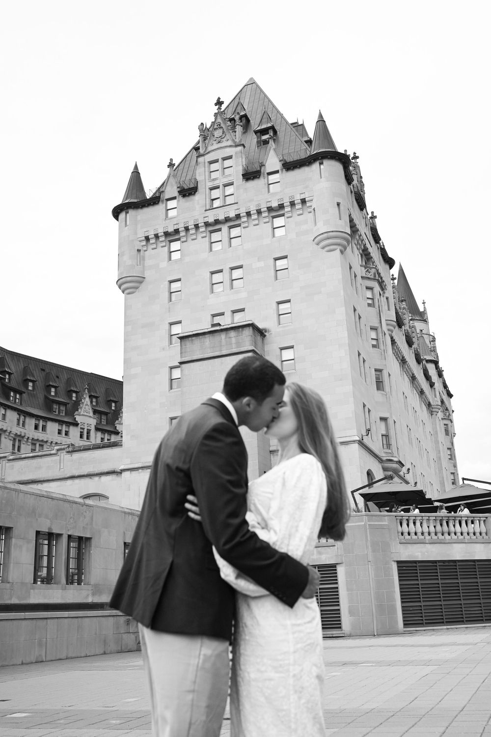 Black and white photo of a couple kissing in front of a grand castle-like building on their wedding day.