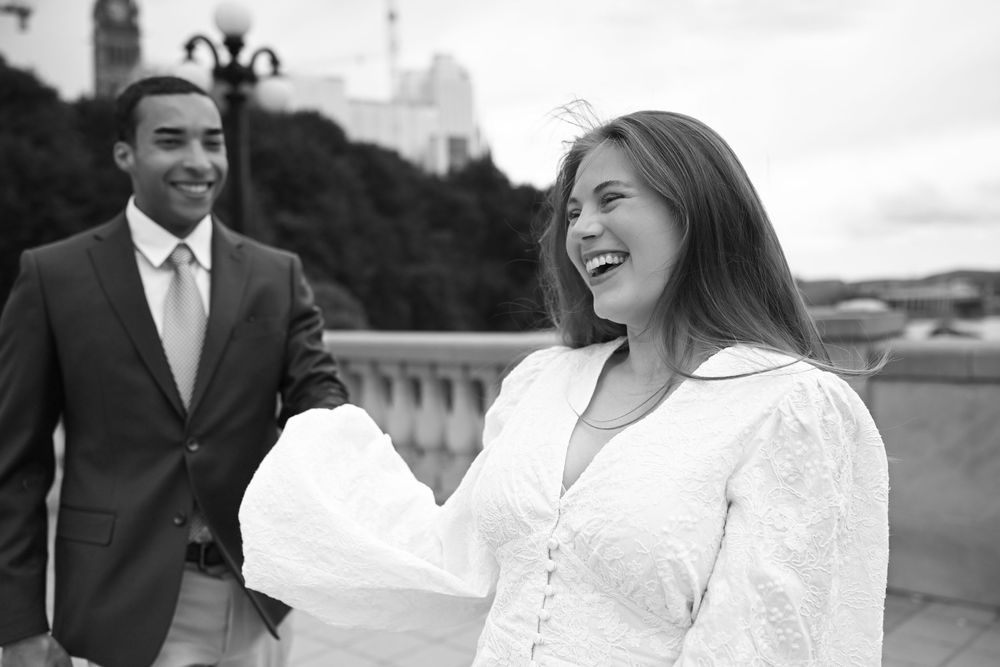 Black and white photo of a laughing woman in white dress with a man in suit on a bridge.
