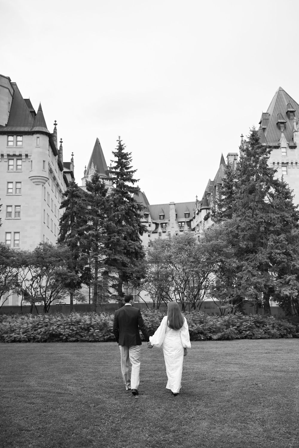 Black and white photo of a couple holding hands walking toward a grand castle-like building surrounded by trees.