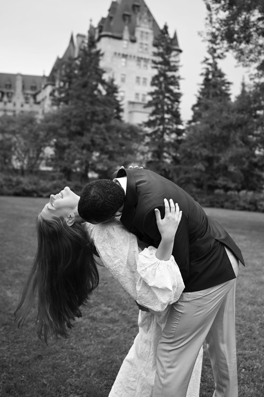 Black and white photo of a couple sharing a romantic dip kiss on a lawn with a grand castle building backdrop.