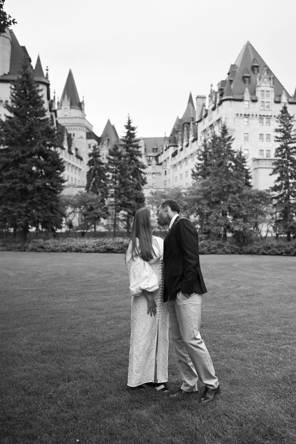 Black and white photo of a couple standing on a lawn in front of a grand château-style building with tall evergreen trees.