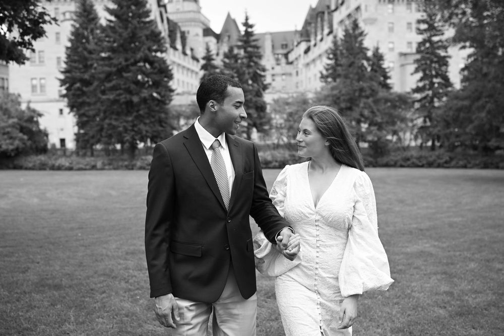 Black and white photo of a couple holding hands and smiling at each other on a green lawn with a grand building behind them.