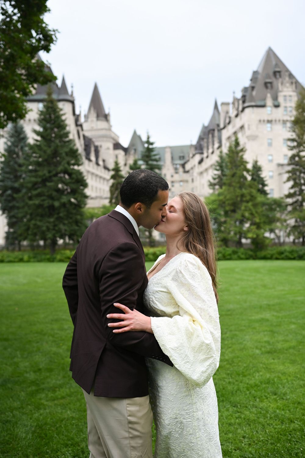 A couple shares a romantic kiss on a green lawn with a grand castle-like building in the background.