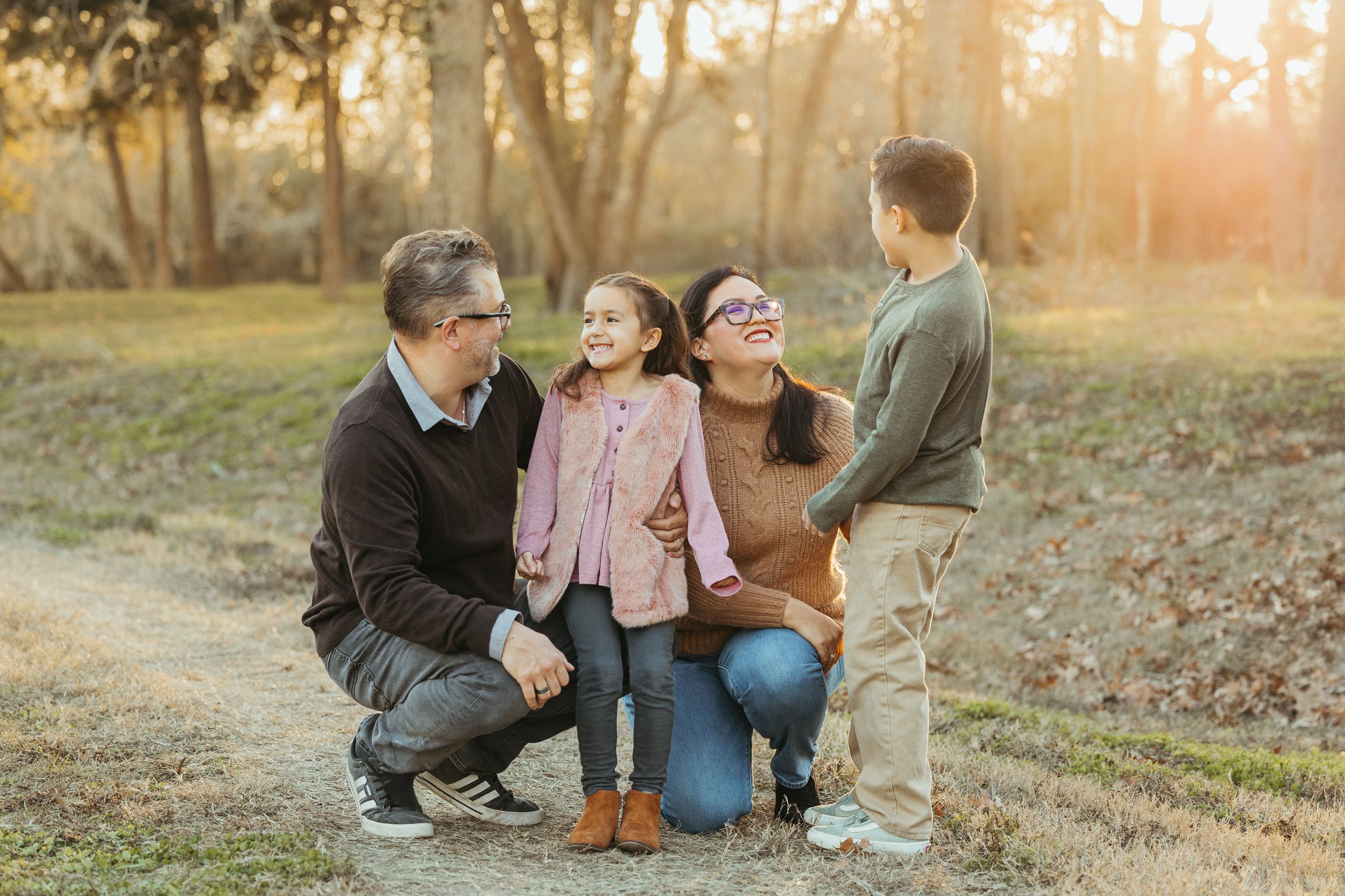 Family of four smiling and laughing together outdoors on a dirt path surrounded by trees in warm golden light.