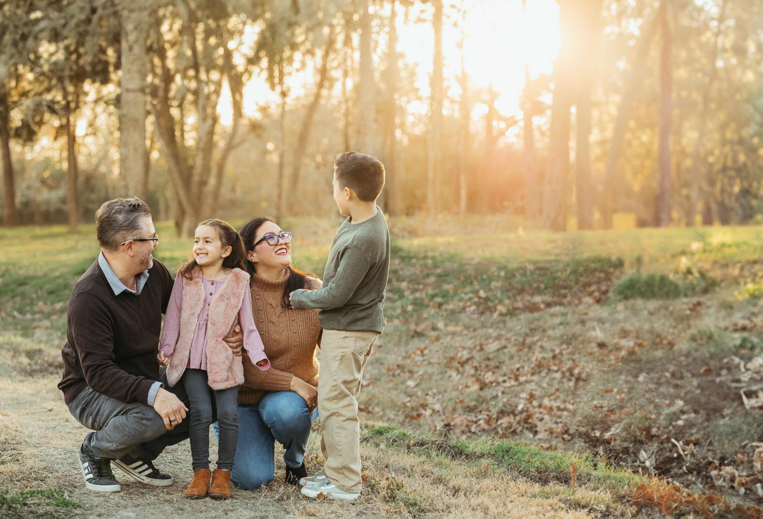 Family of four sharing a warm moment outdoors at golden hour in a wooded park setting.