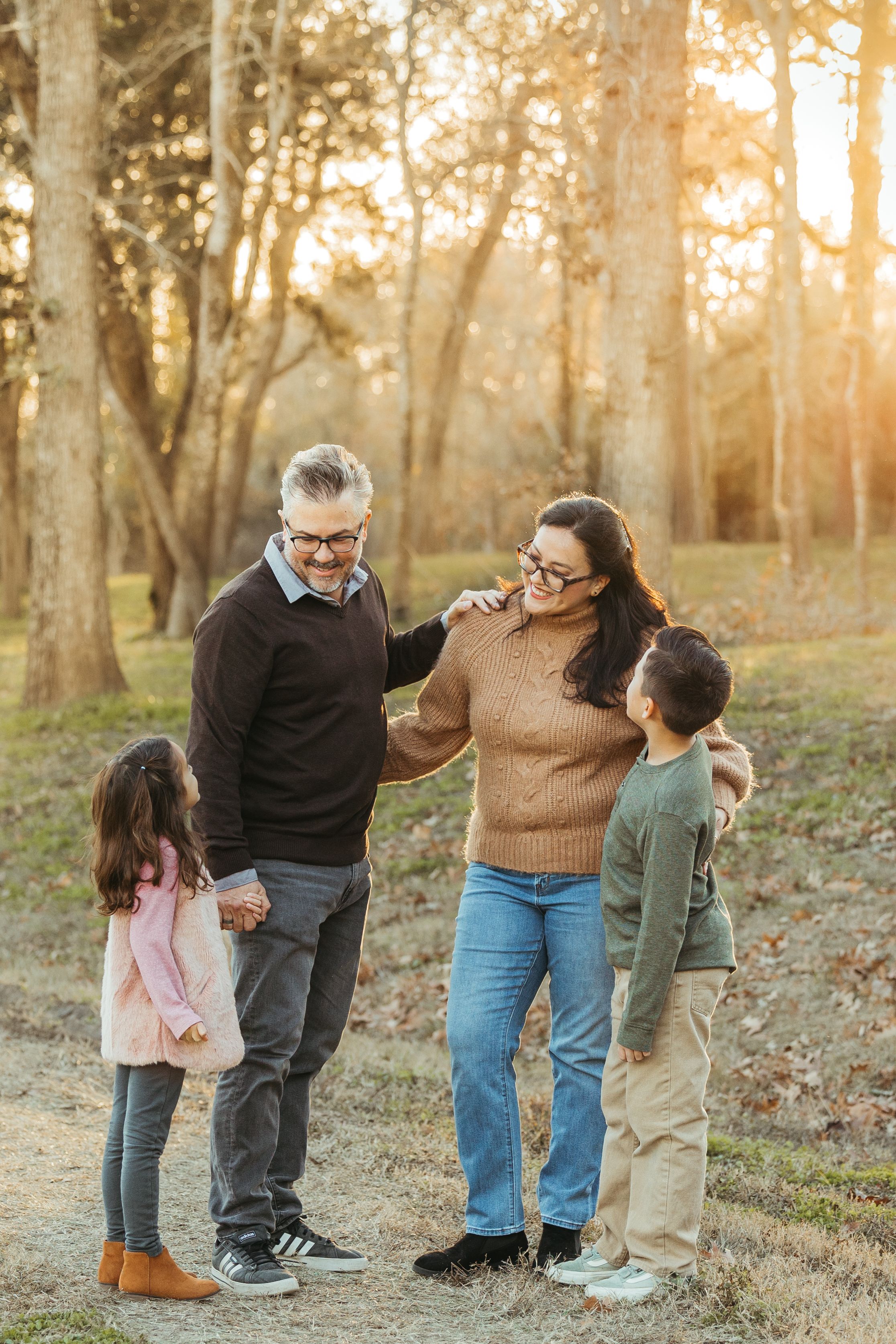 Family of four smiling together in a sunlit autumn park, parents with two young children enjoying a warm outdoor moment.