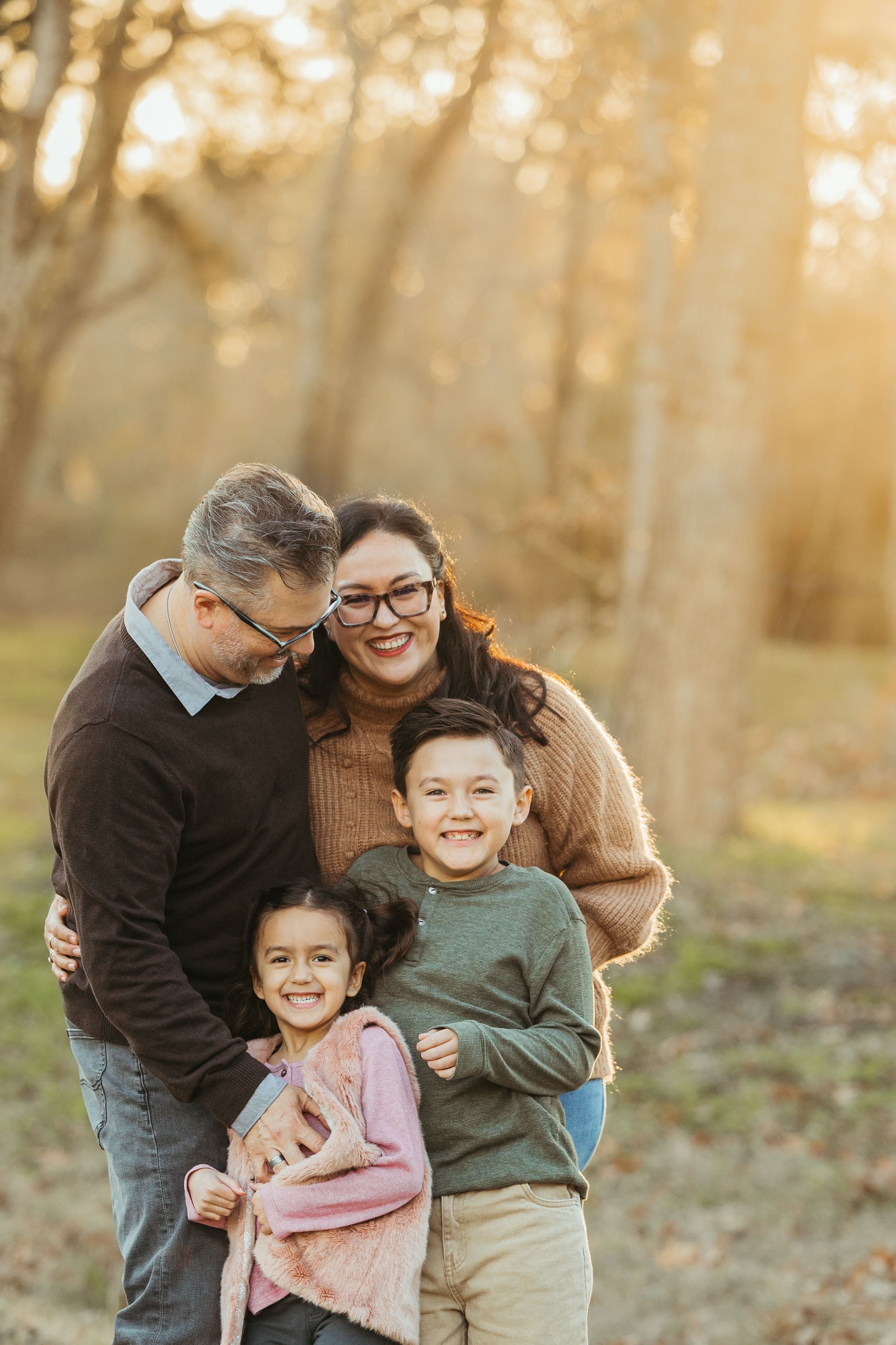 A happy family of four poses outdoors in golden autumn light, with parents embracing their smiling son and daughter.