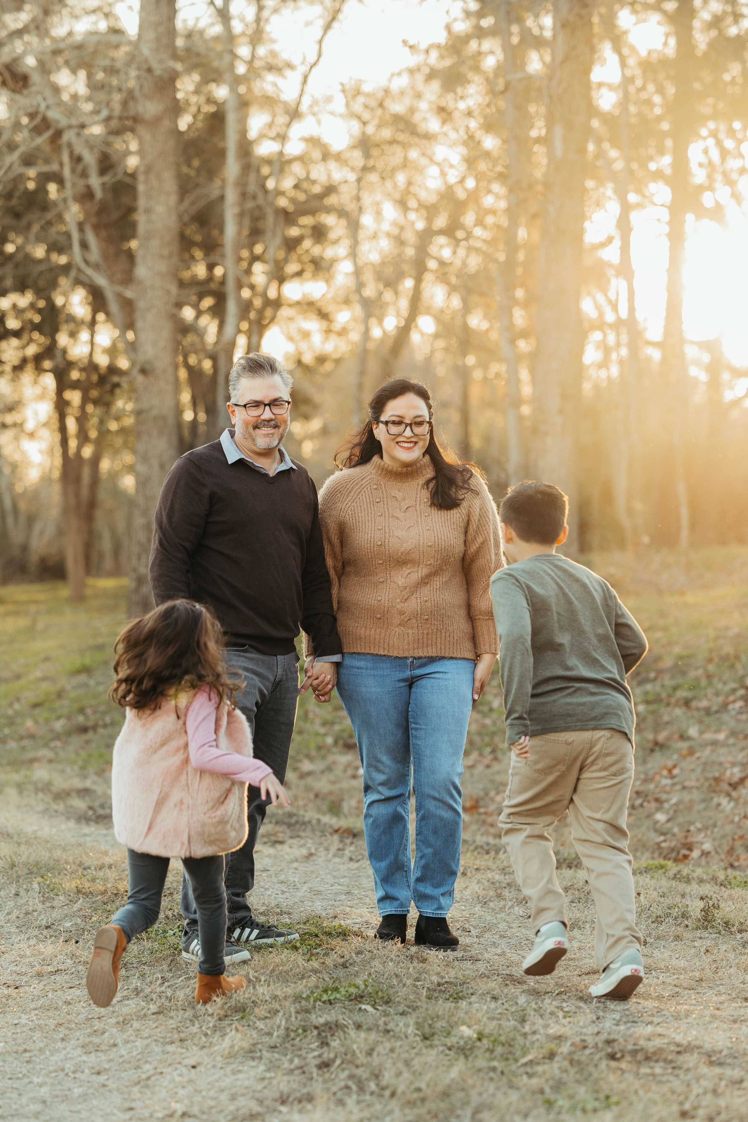 Family of four walking hand-in-hand on a sunlit forest path during golden hour.