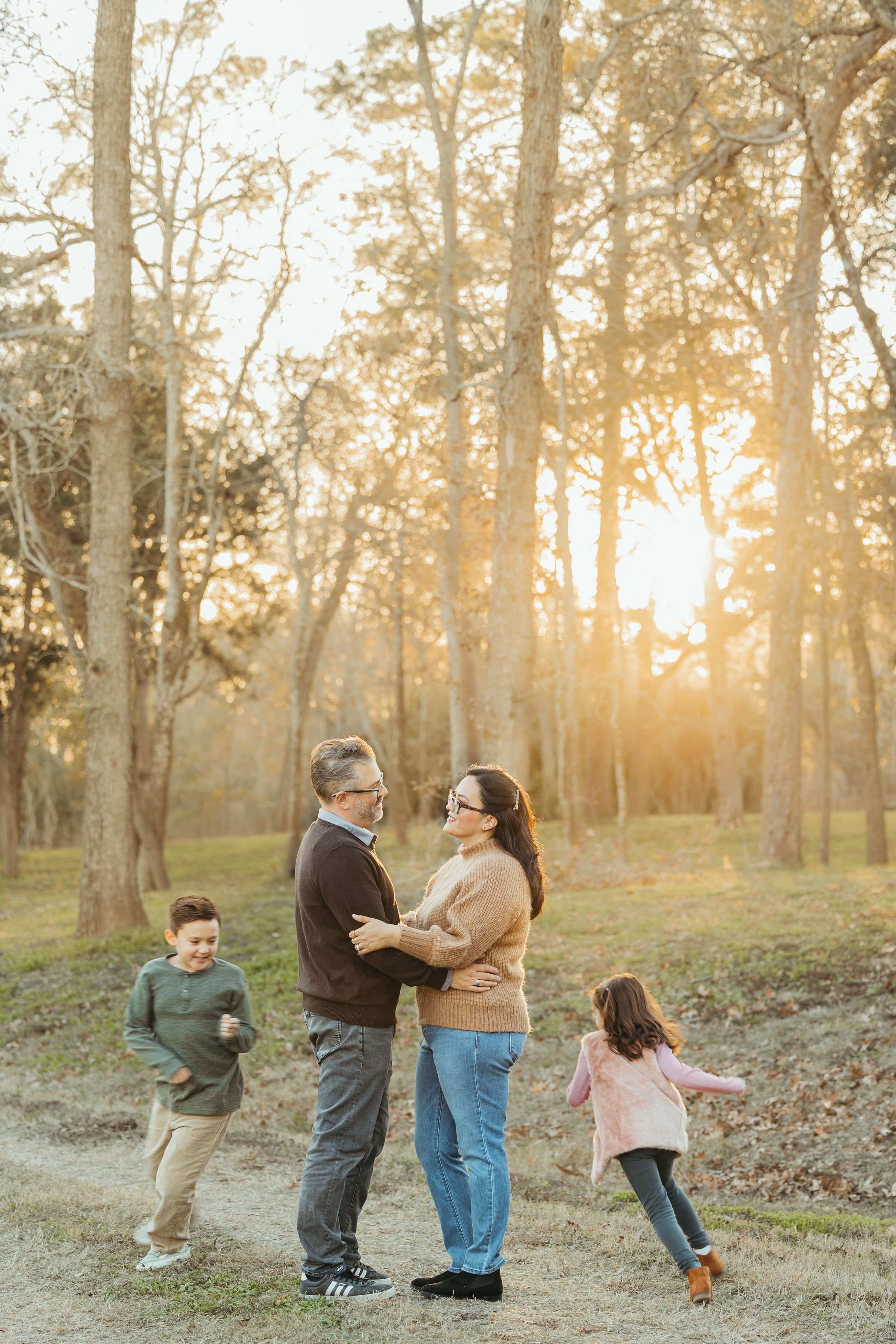 Family of four enjoying a golden hour sunset in a wooded park, parents embracing while children play around them.