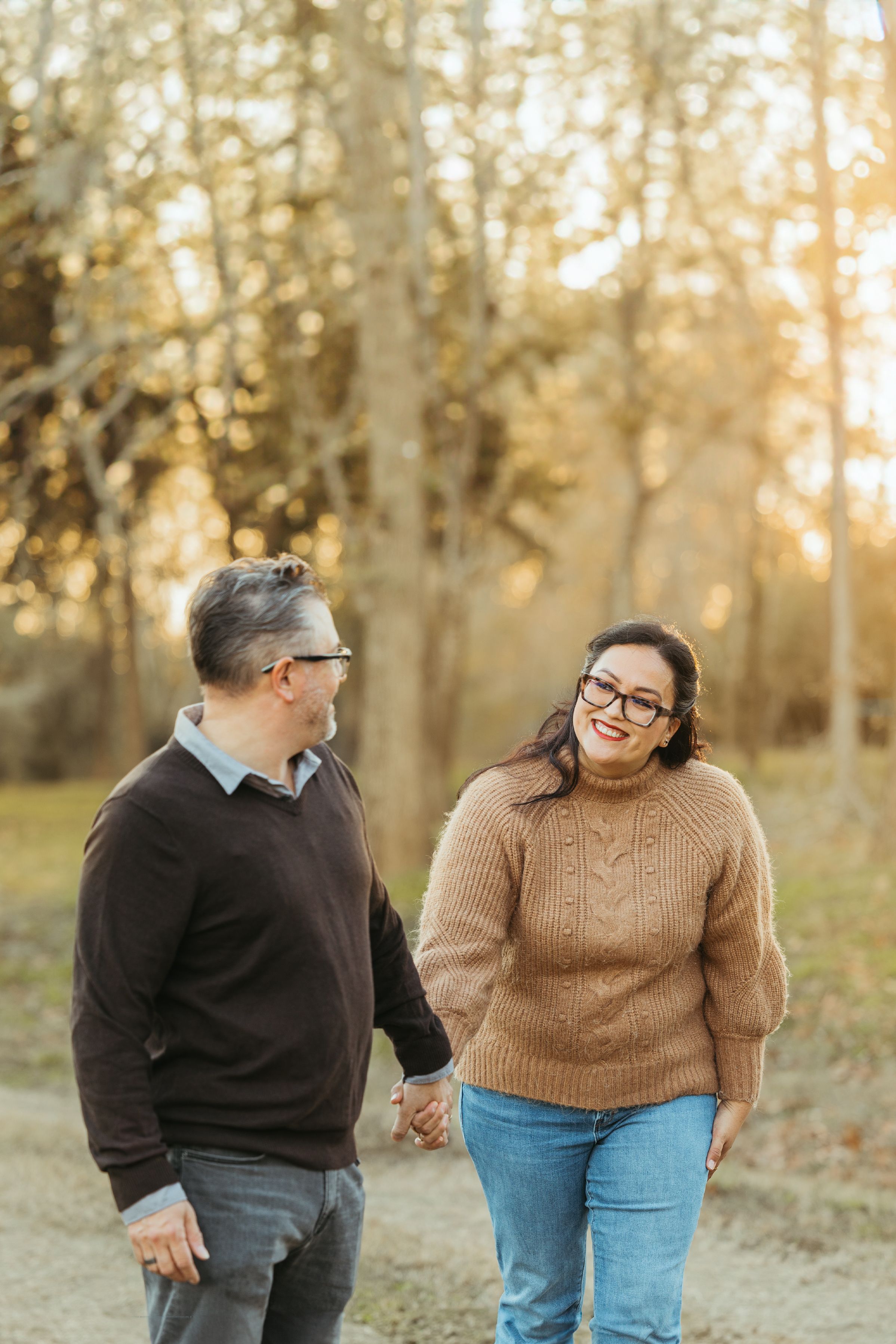 Couple holding hands and smiling at each other while walking outdoors in a sunlit autumn park.