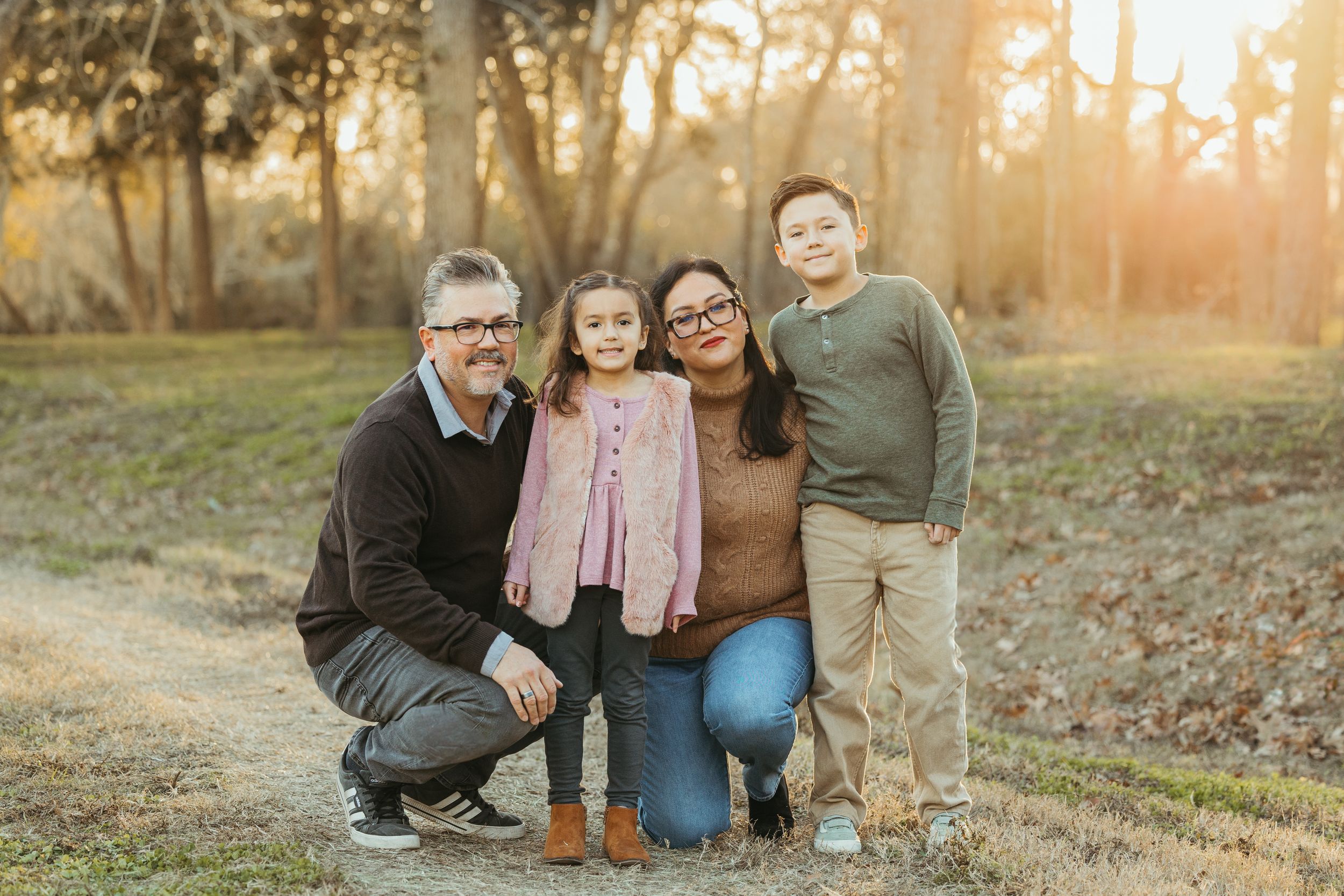 Family of four posing together on a sunlit forest path during golden hour outdoor photo session.