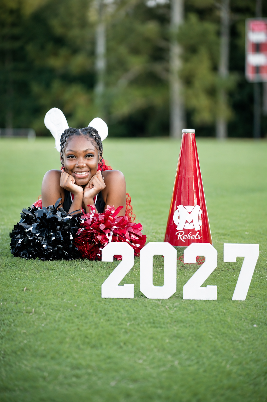 A cheerleader poses on a football field with pom-poms, a red Rebels megaphone, and a 2027 sign.