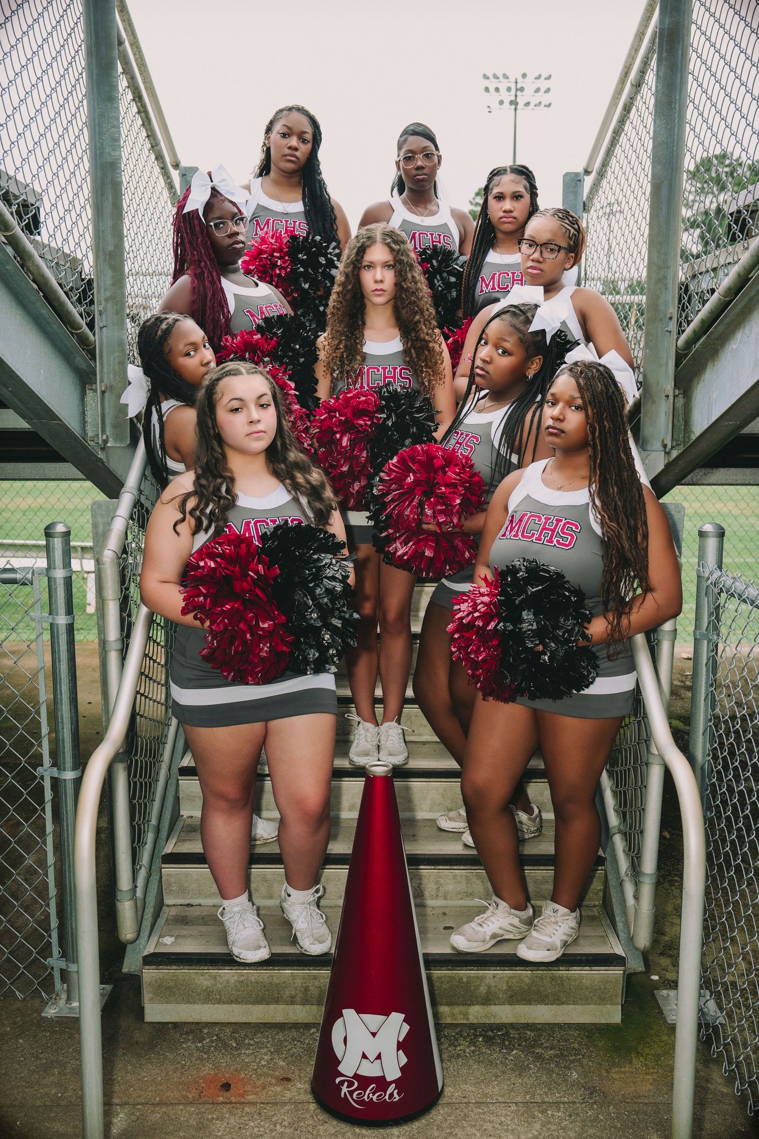 High school cheerleading squad poses on stadium stairs with red and black pom-poms and a Rebels megaphone.