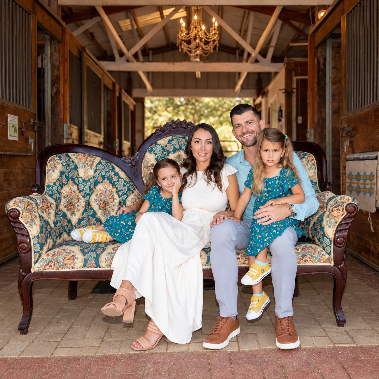 Family of four smiling on a floral sofa in a rustic barn, parents with two young girls in matching teal outfits.