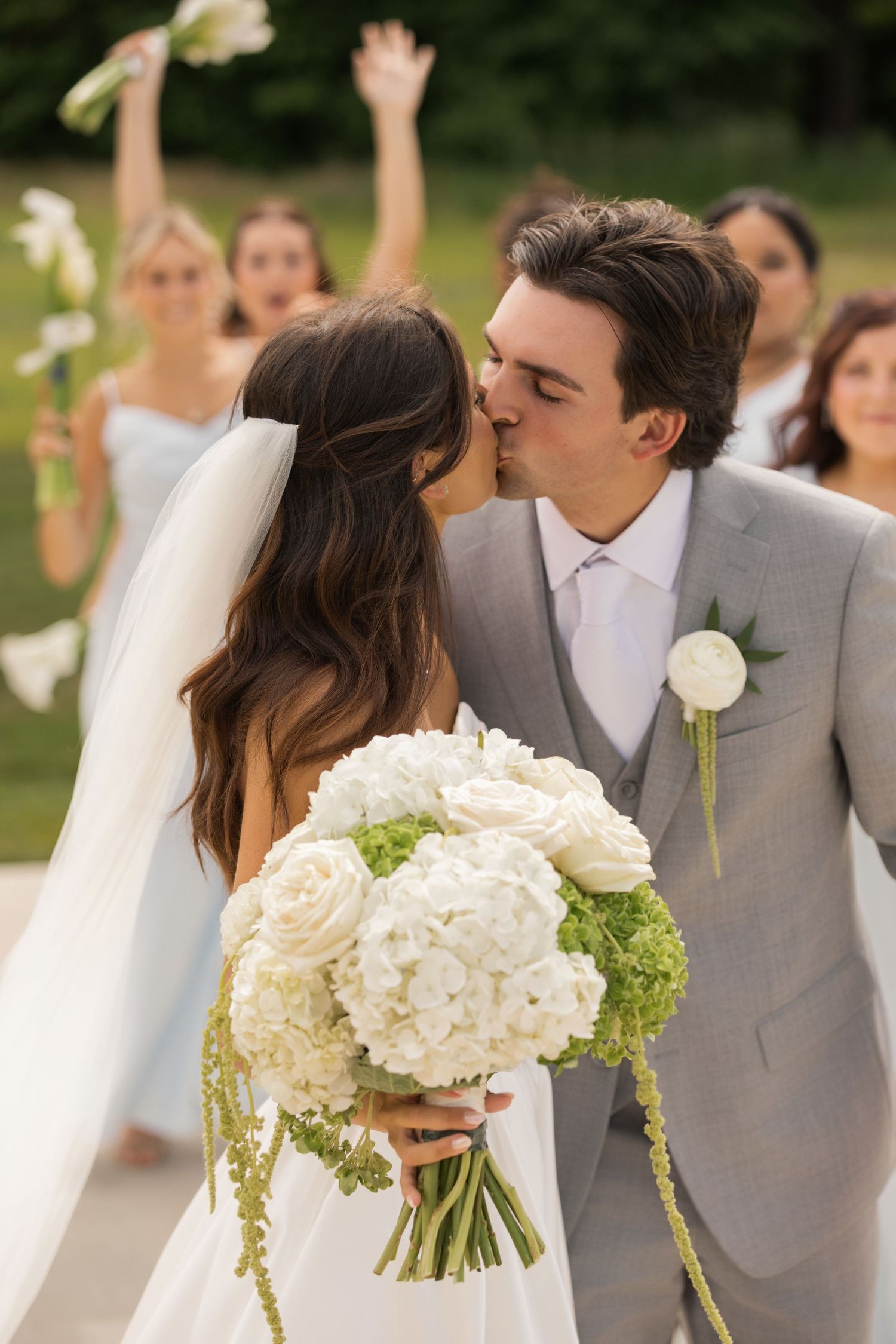 Bride and groom share a kiss at outdoor wedding ceremony, bride holding white hydrangea bouquet, bridesmaids celebrating behind them.