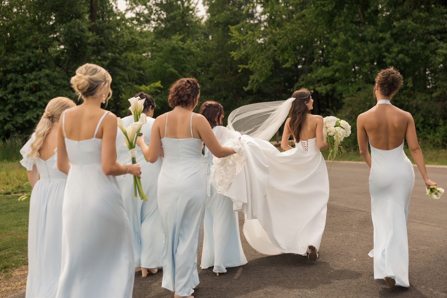 Bride and bridesmaids in light blue gowns walking together outdoors, holding white bouquets with lush green trees in background.