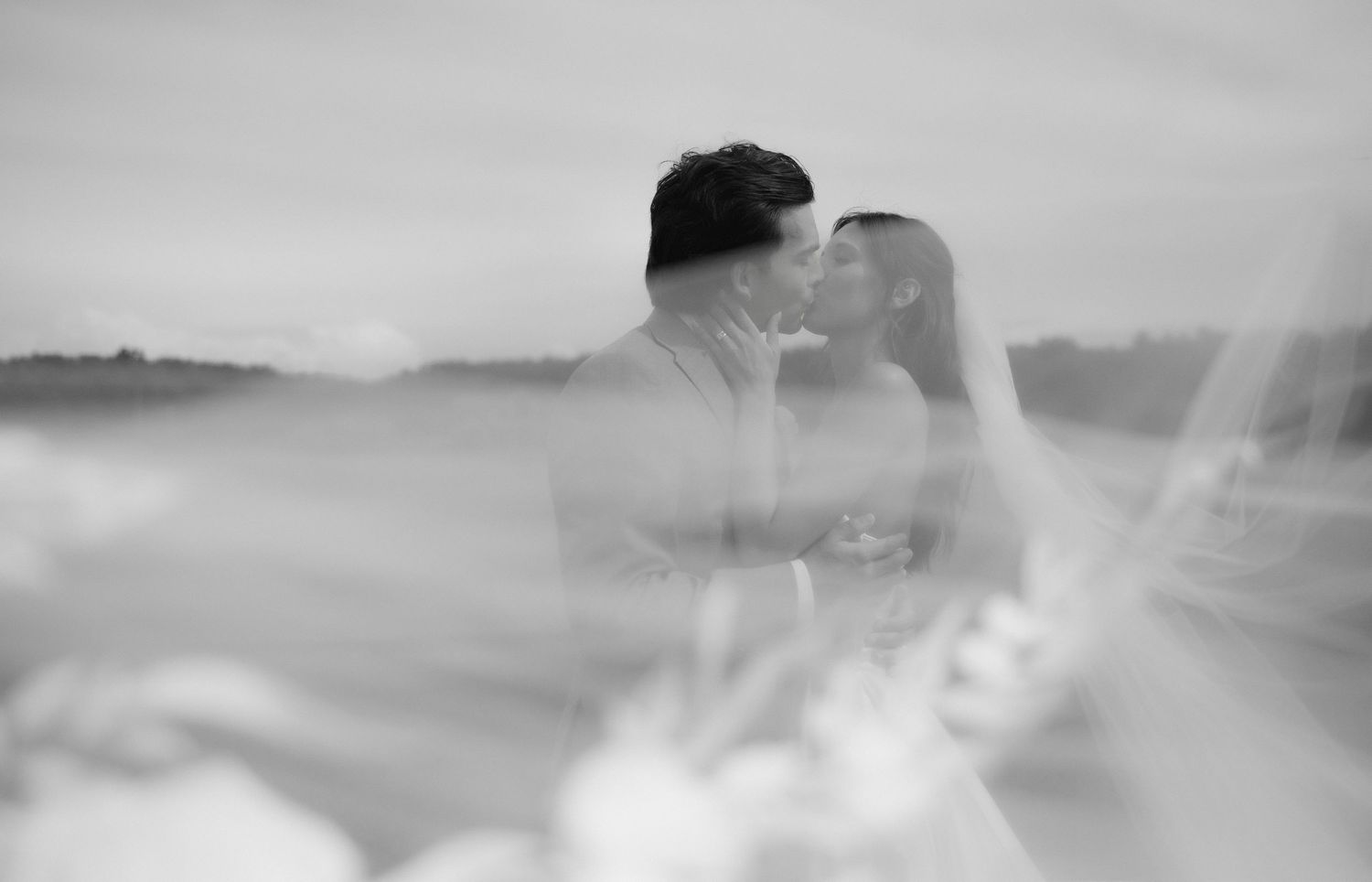 Black and white wedding photo of bride and groom sharing a romantic kiss outdoors, with soft bokeh background.