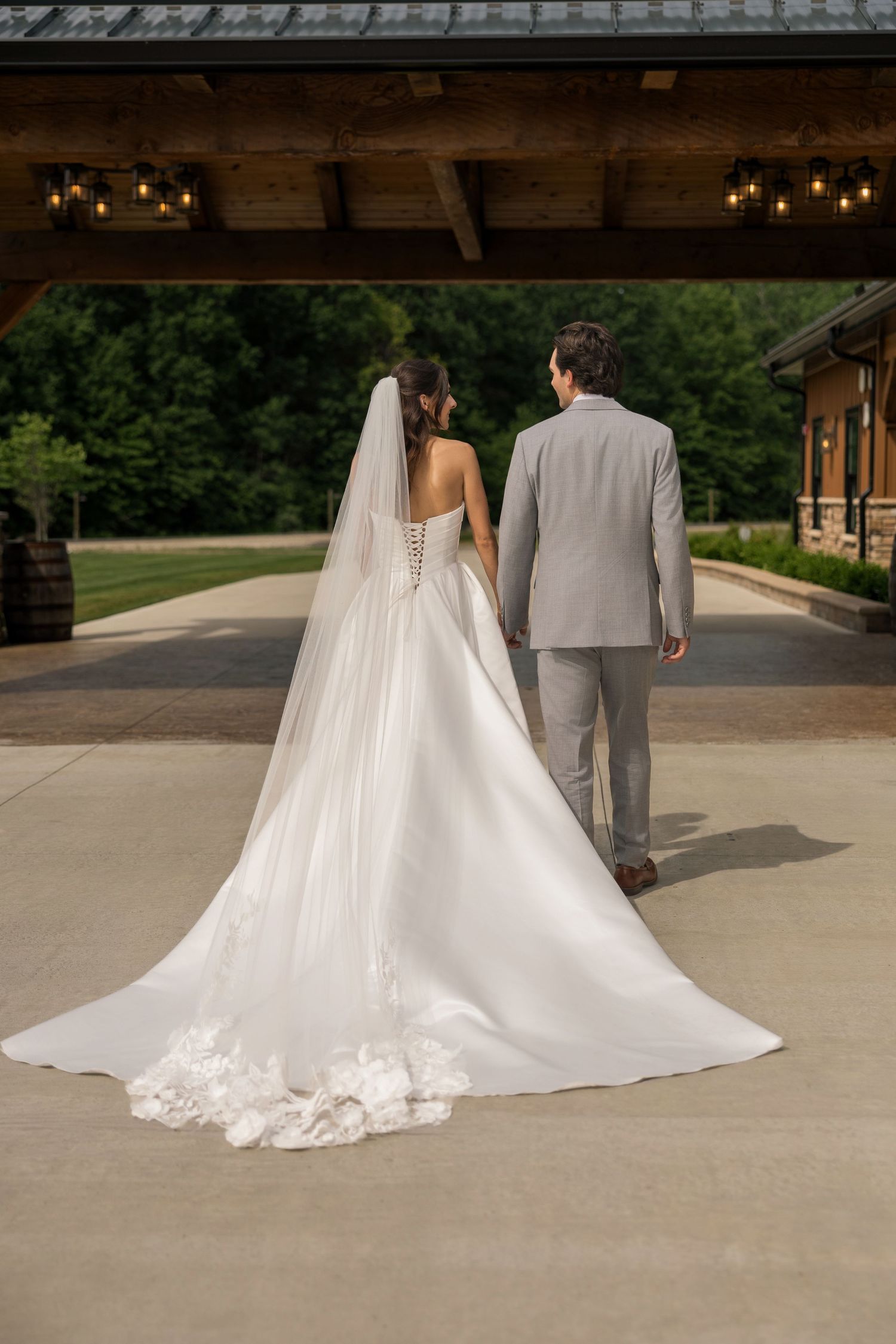 Bride in white gown with long veil and groom in gray suit walk hand in hand at elegant outdoor wedding venue.