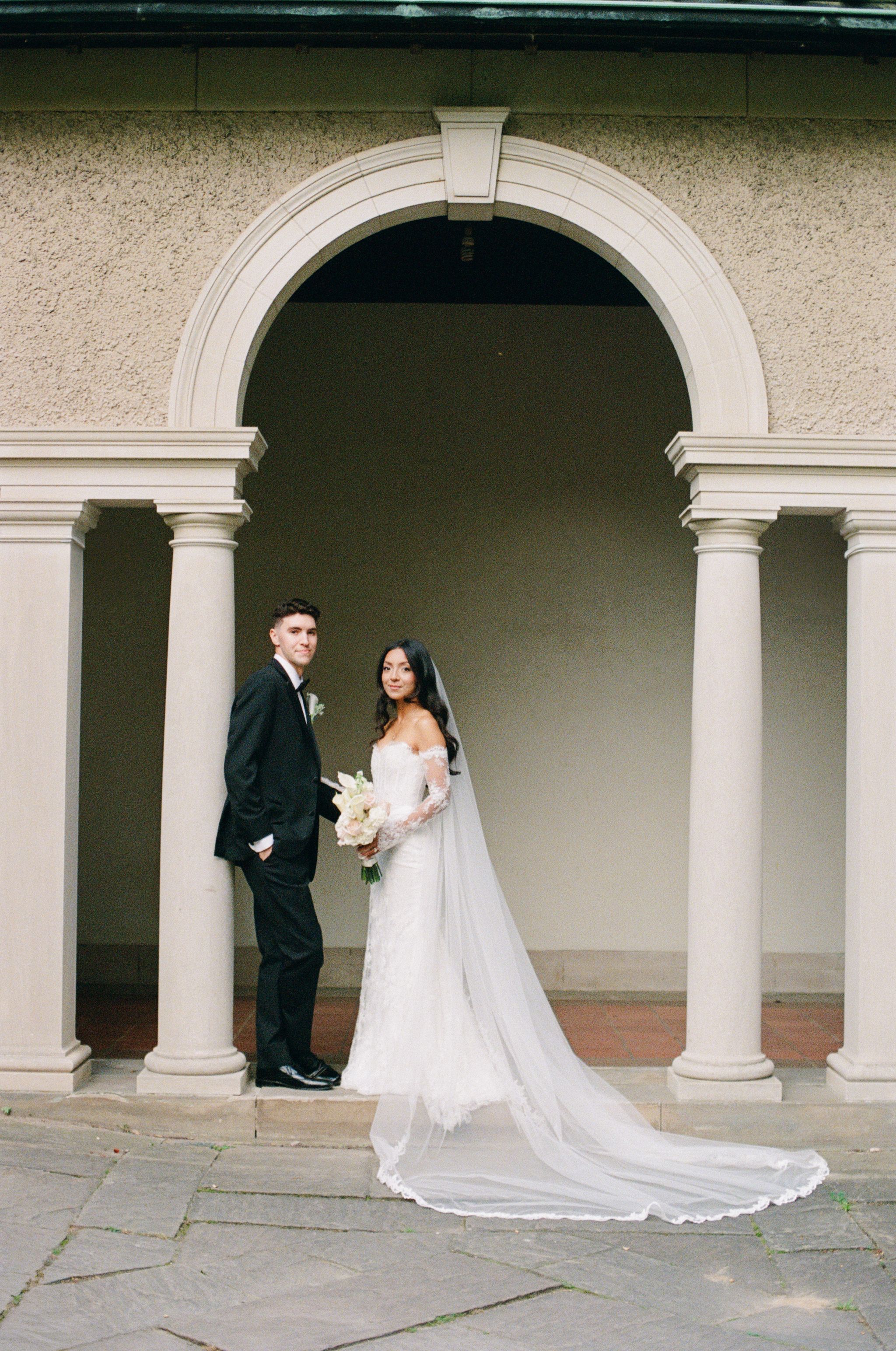Bride in a white lace gown with a long veil and groom in a black suit pose together beneath a stone archway with columns.