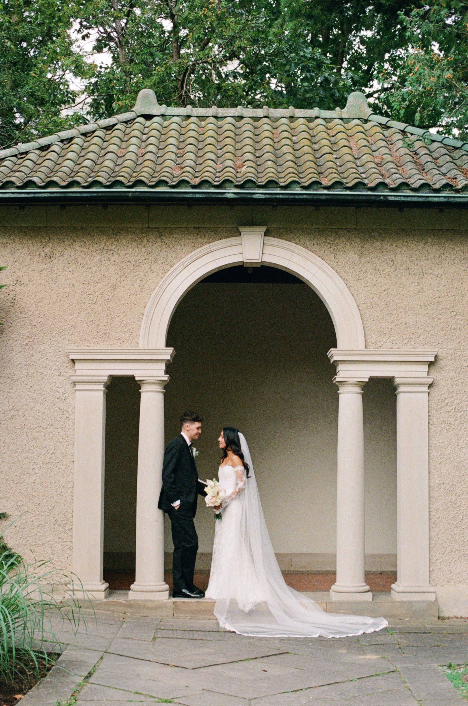 Bride in a long veil and groom in a black suit pose together beneath a stone archway with columns at a terracotta-roofed garden pavilion.