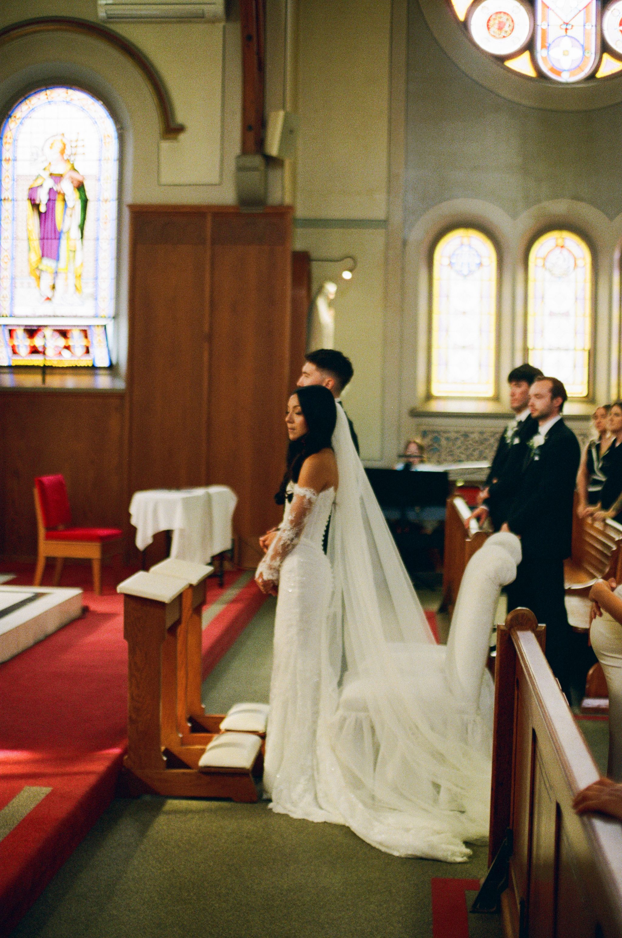 Bride in a long lace gown and veil stands beside her groom at the altar of a church with stained glass windows and guests looking on.