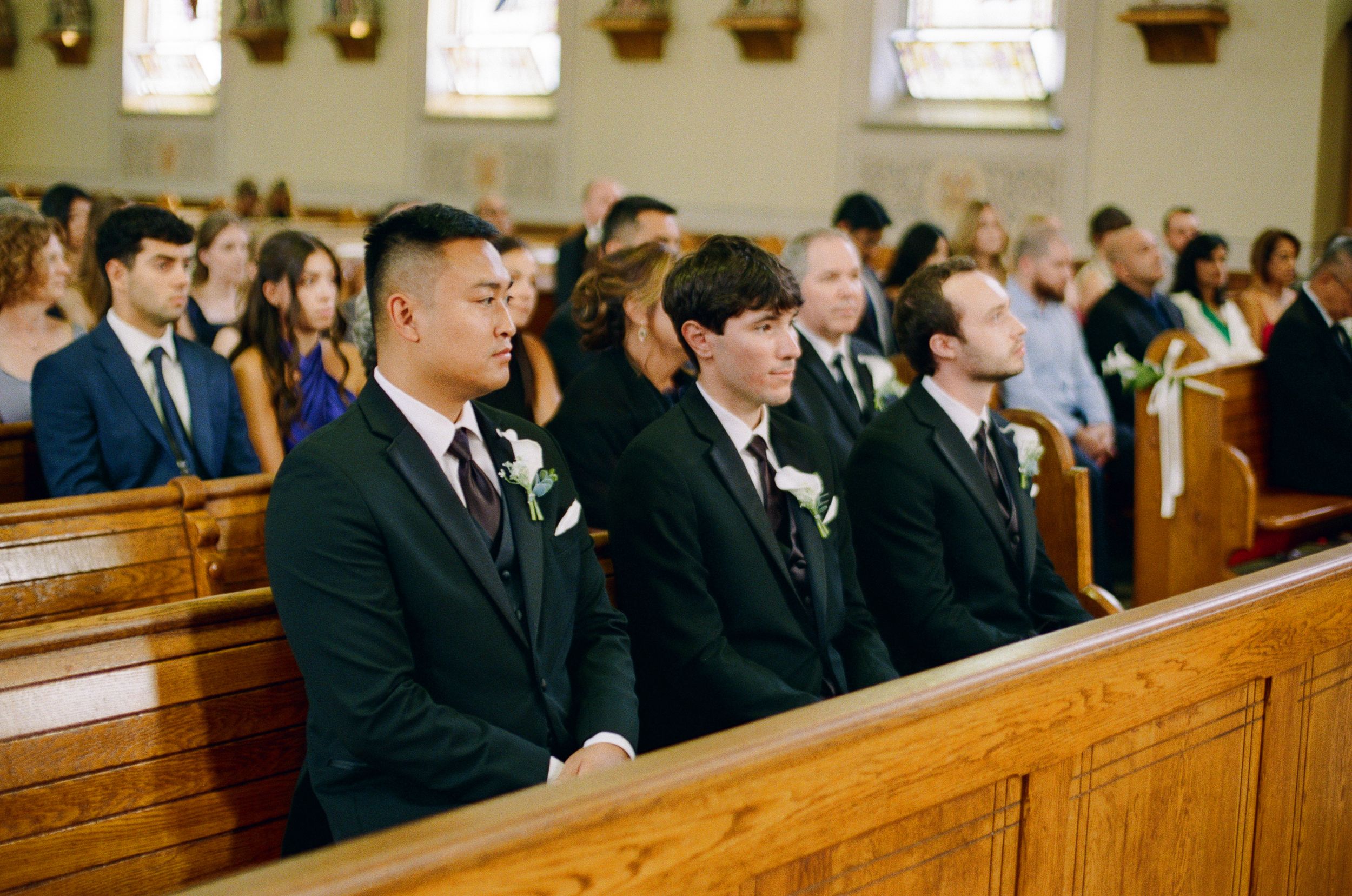 Three groomsmen in dark suits with white floral boutonnieres stand in front of seated wedding guests inside a church.