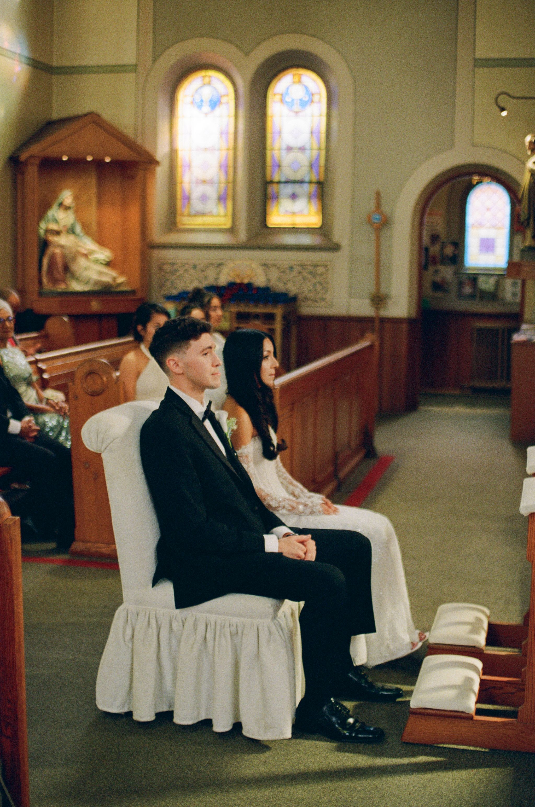 Bride in a white lace gown and groom in a black tuxedo sit together on white-draped chairs during a church wedding ceremony.