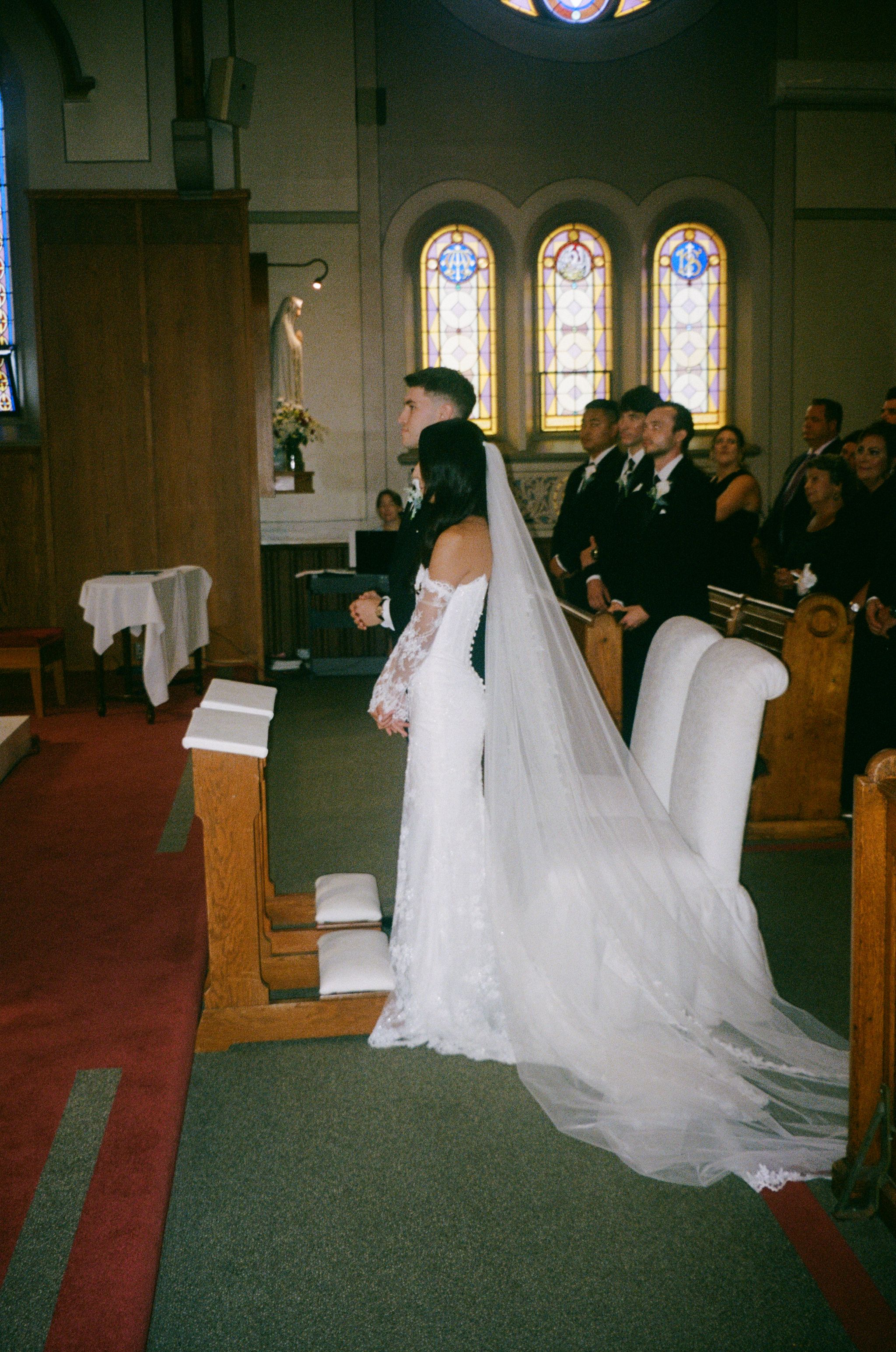 Bride in a lace gown with a long cathedral veil stands beside the groom at the altar of a church as wedding guests look on.