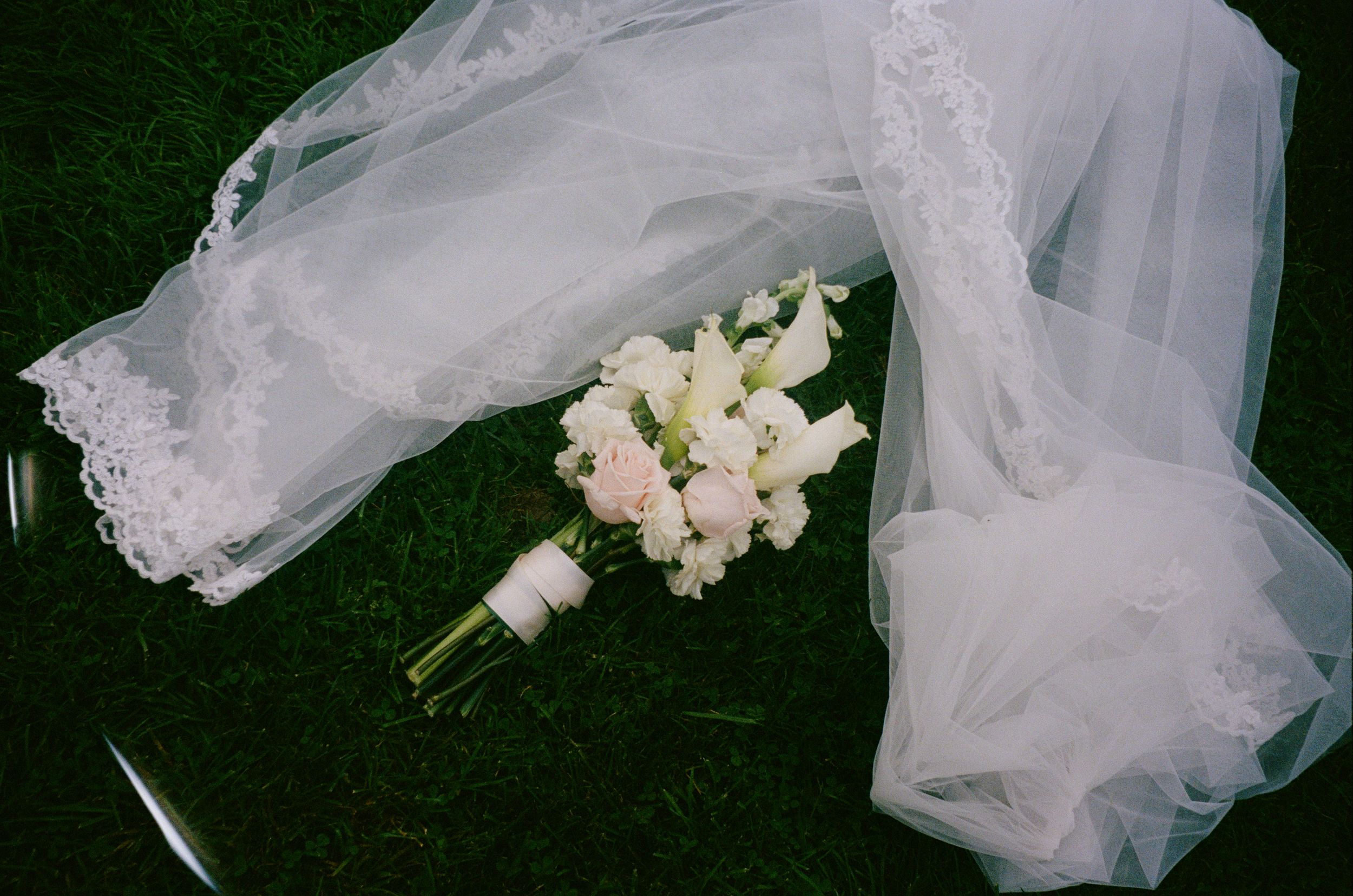 White bridal bouquet of roses and lilies tied with ribbon resting on green grass beside a lace-trimmed wedding veil.