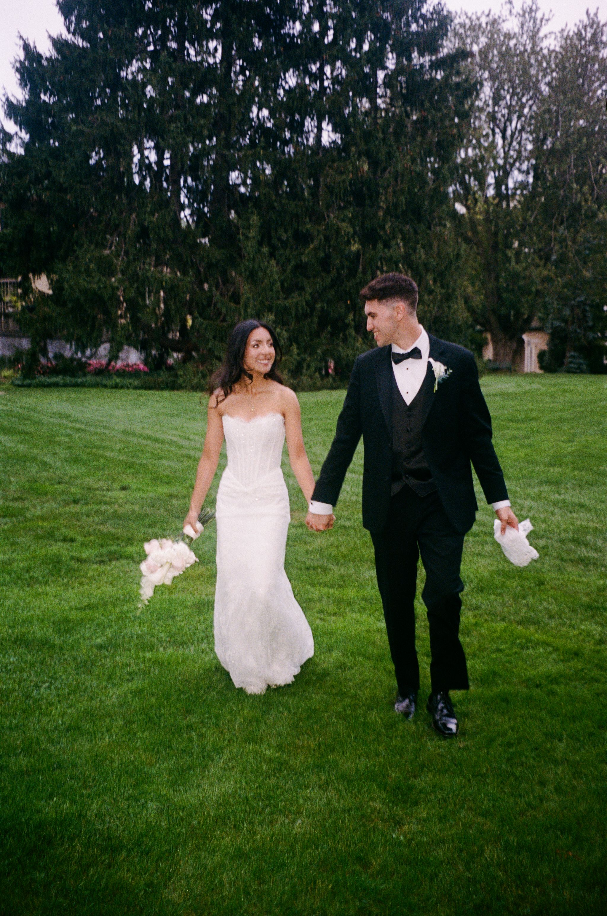 Bride in a strapless white gown and groom in a black tuxedo hold hands while walking across a green lawn with tall evergreen trees behind.