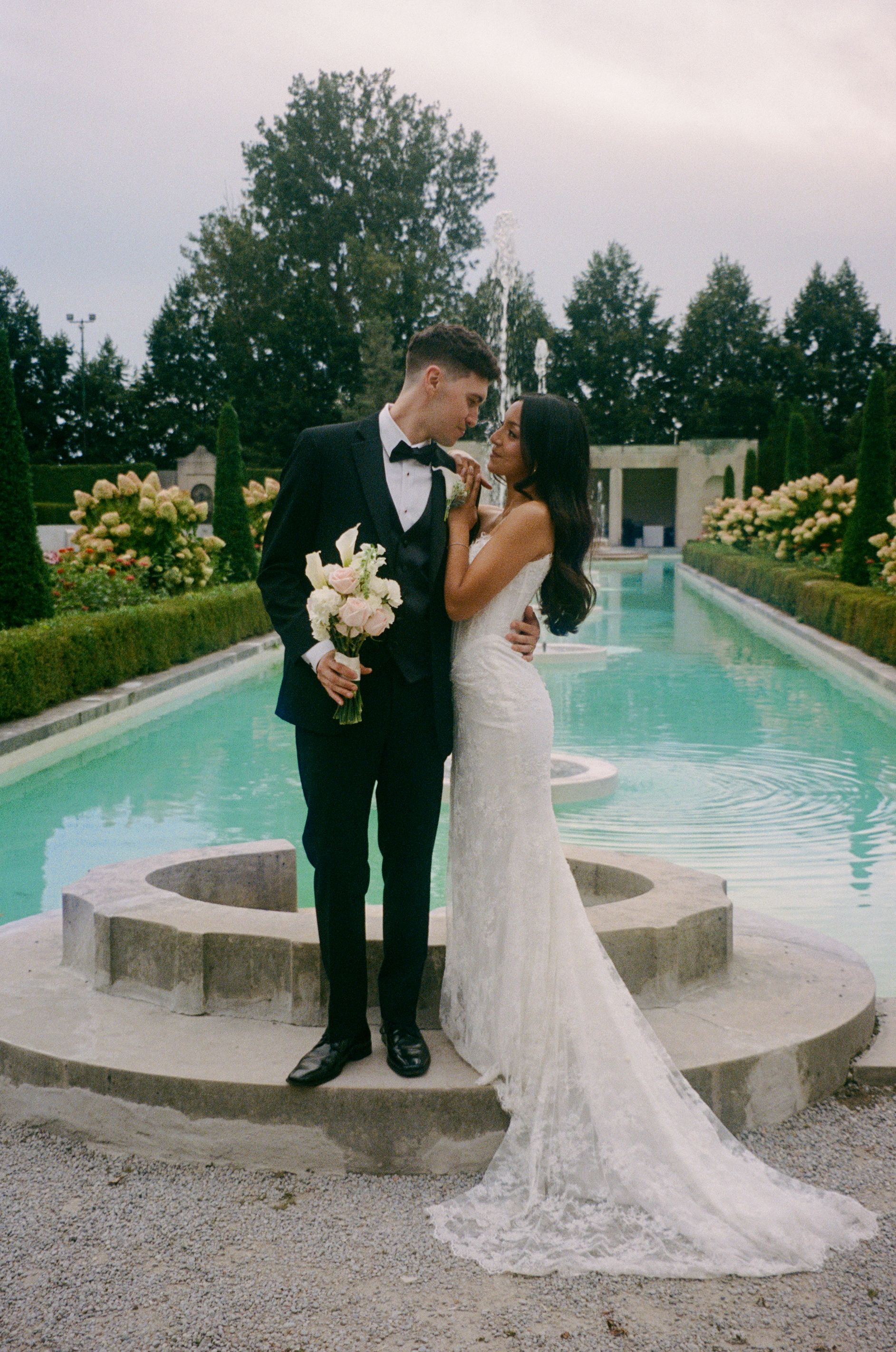 Bride in a lace wedding gown and groom in a black tuxedo holding a pink bouquet embrace beside a reflecting pool in a lush garden.