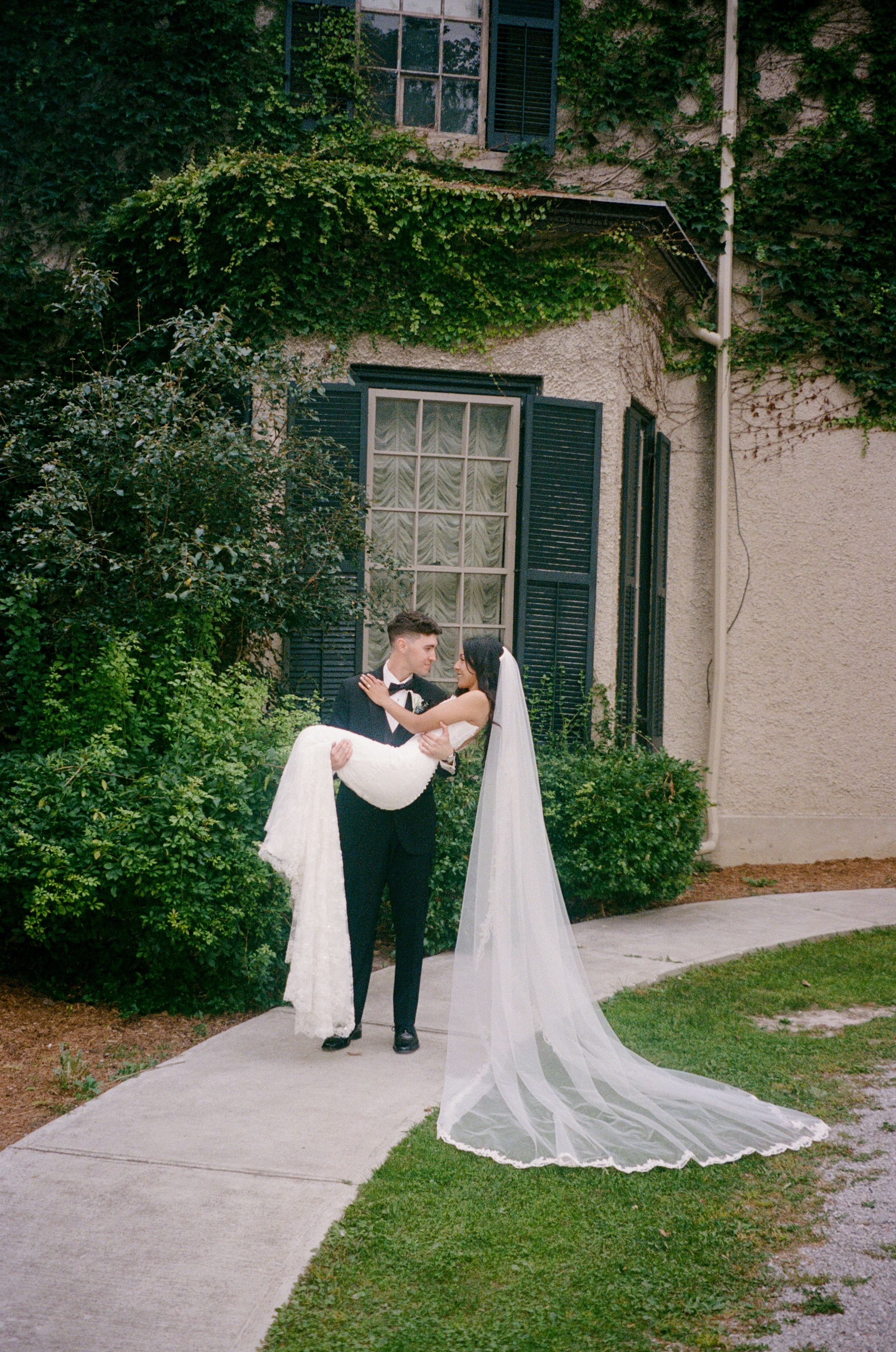 Groom in a black tuxedo carries his bride in a white gown with a long veil along a garden path beside an ivy-covered stone building.