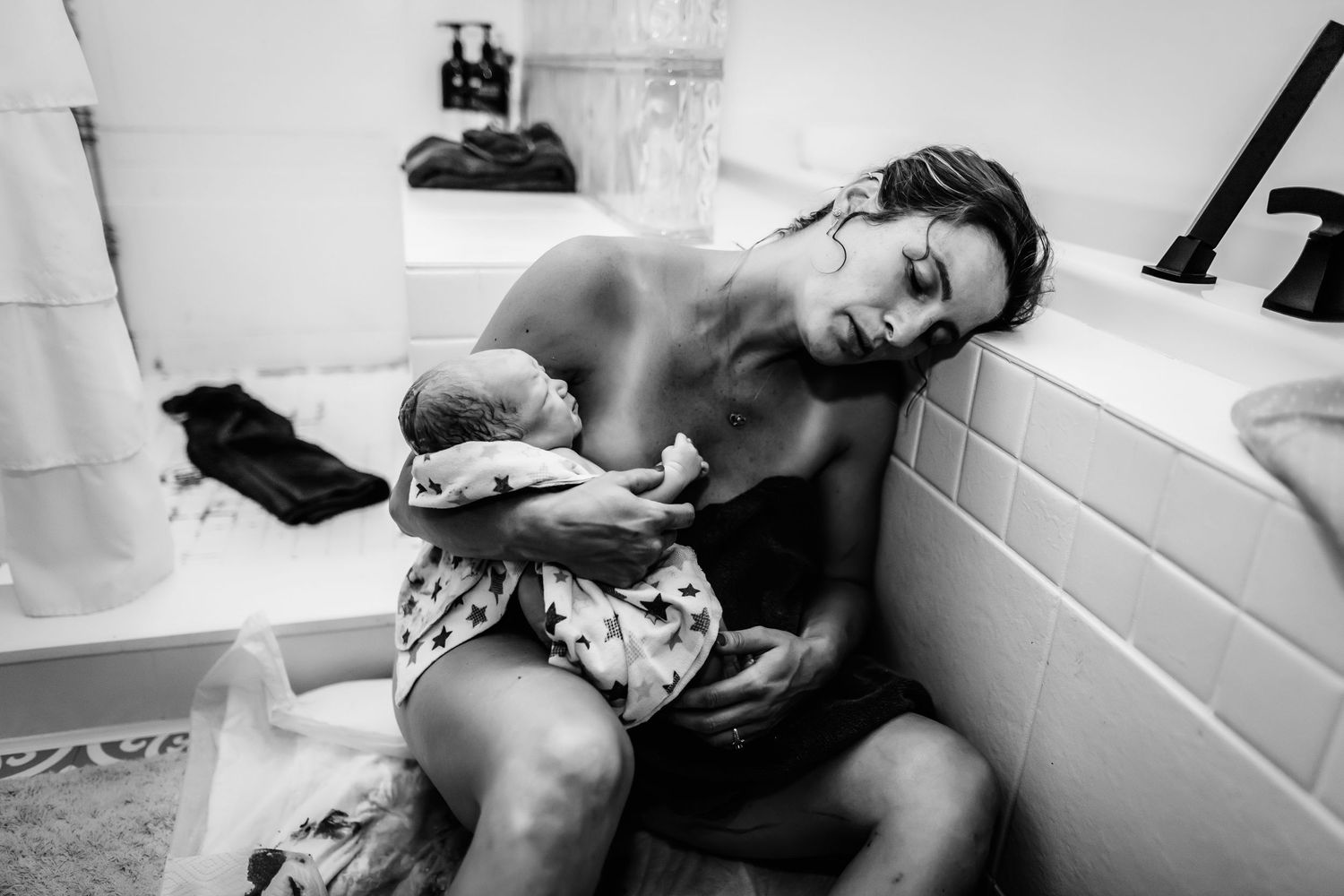 Black and white photo of a mother sitting beside a bathtub, cradling her newborn baby wrapped in a blanket after a home birth.