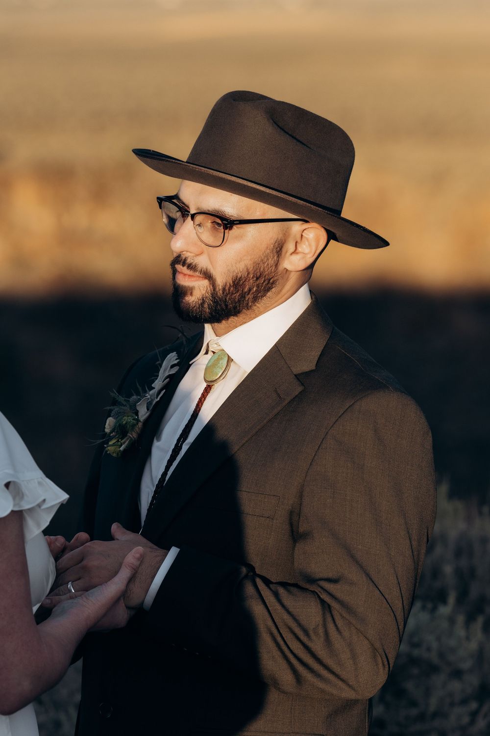 Bearded groom in brown suit, wide-brim hat, and glasses holds hands during a golden hour desert wedding ceremony.