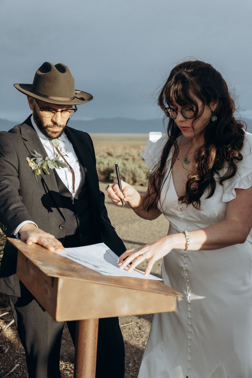 Bride in white dress signs marriage license at wooden podium while groom in suit and hat watches outdoors.
