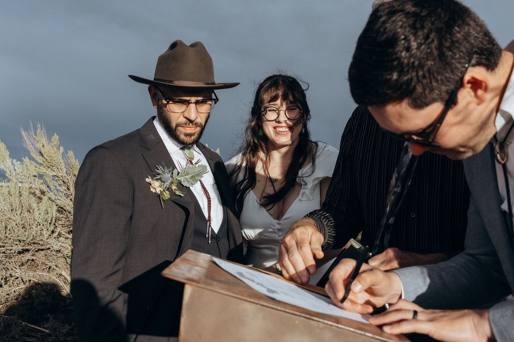 Couple signs wedding documents outdoors with officiant, groom wearing brown hat and boutonniere, bride in glasses smiling.