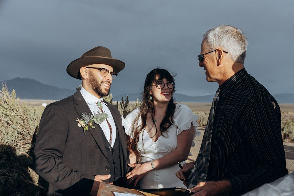 Couple exchanges vows outdoors with officiant, groom in brown hat and suit, bride in white dress, mountains in background.