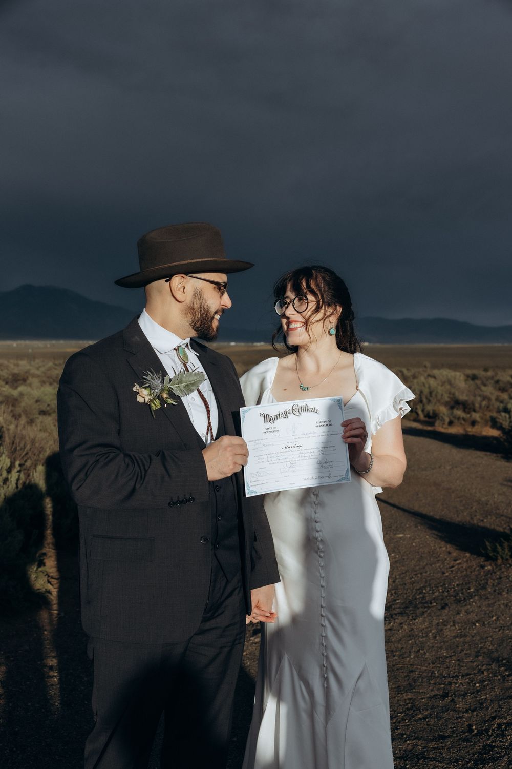 Couple in wedding attire holding marriage certificate outdoors under dramatic stormy sky in desert landscape.