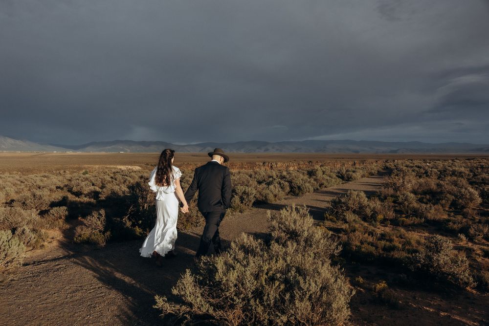 A bride and groom walk hand in hand through a desert landscape under dramatic storm clouds.