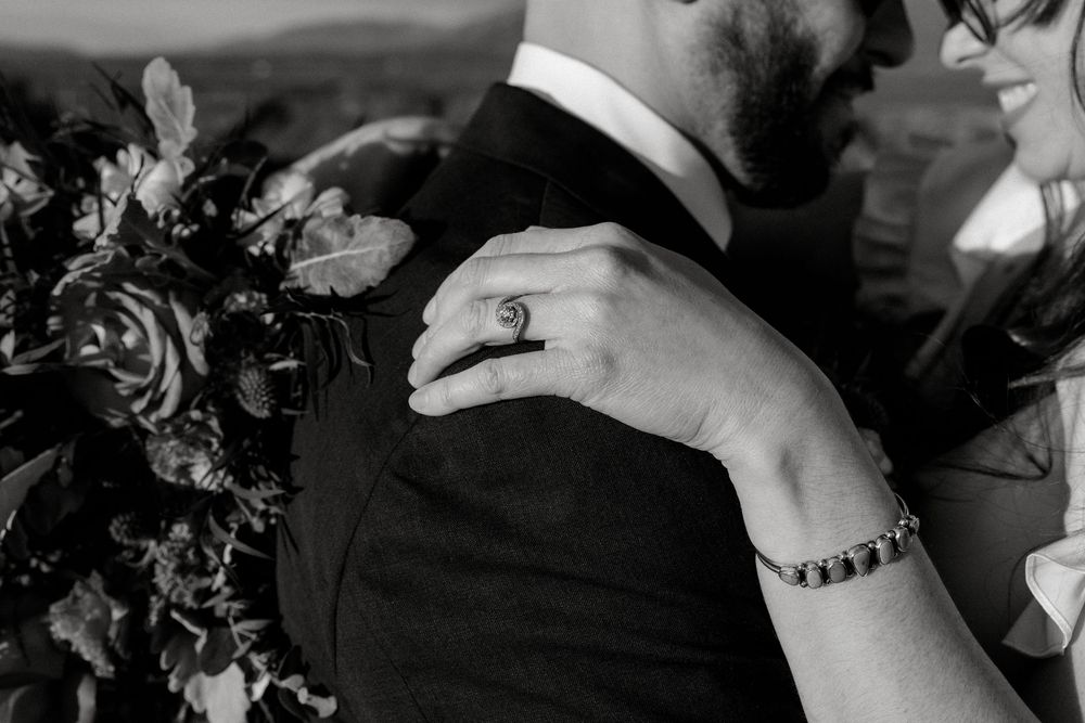 Black and white wedding photo of bride's hand with ring and bracelet resting on groom's shoulder near floral bouquet.