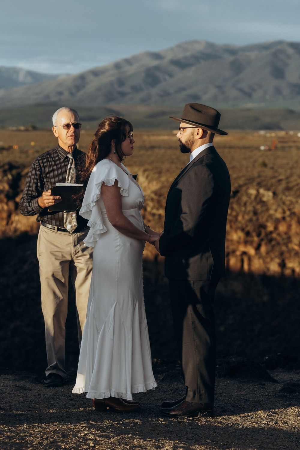 Couple exchanges vows outdoors at golden hour with officiant, mountains backdrop, bride in white dress, groom in dark suit and hat.
