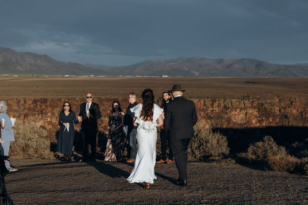 Outdoor wedding ceremony in a desert landscape with mountains, guests watching couple exchange vows under dramatic cloudy skies.