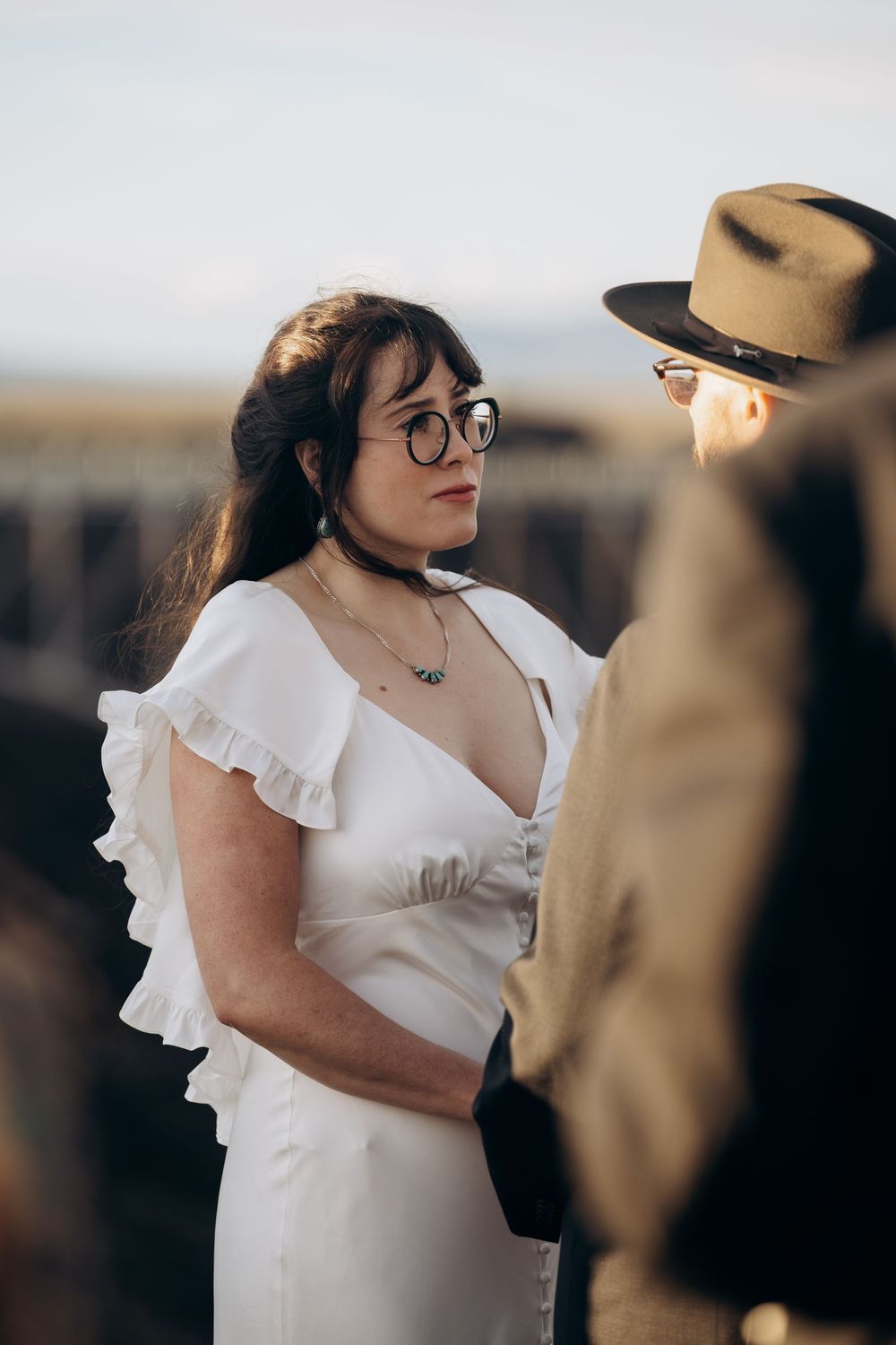 Bride in white ruffled dress and glasses exchanges vows outdoors with partner in vintage hat.