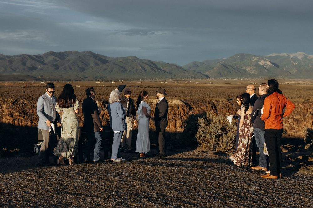 Small group of people gathered outdoors at sunset with vast desert plains and mountain range backdrop.
