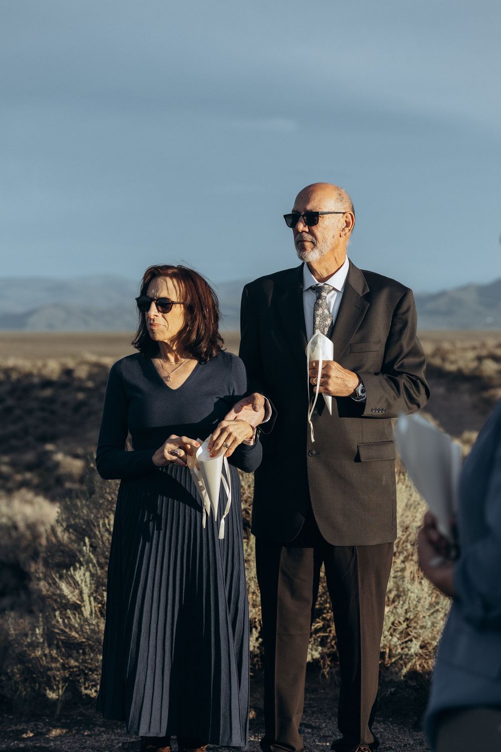 Elegantly dressed couple in sunglasses holding white masks at an outdoor desert wedding ceremony.