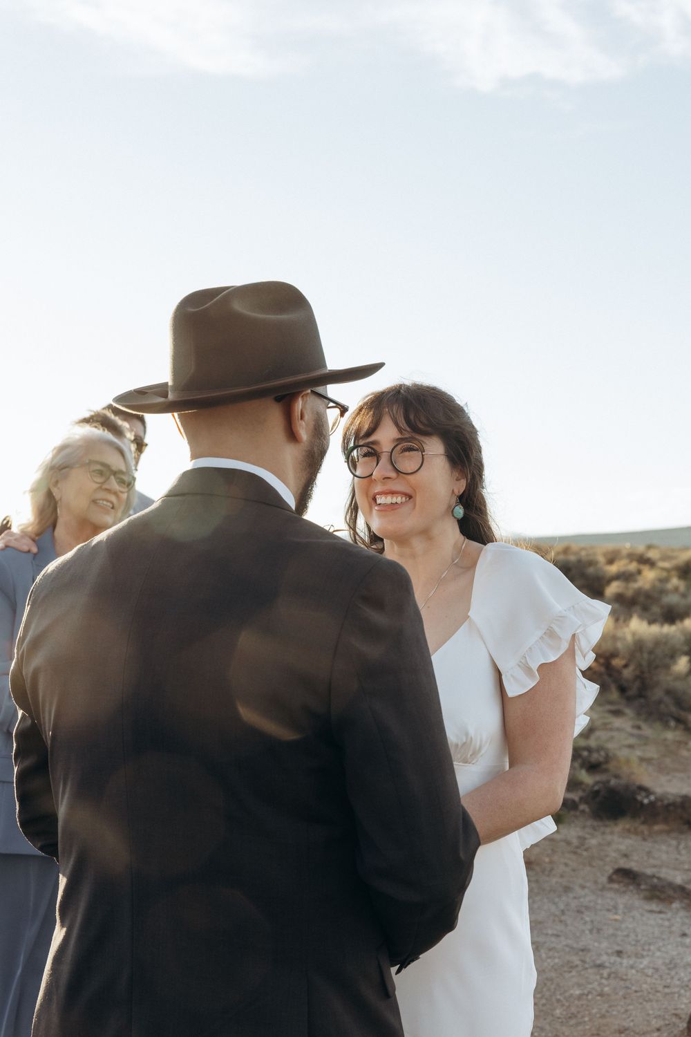 Smiling bride in white dress with glasses holds hands with groom in brown suit and hat during outdoor desert wedding ceremony.
