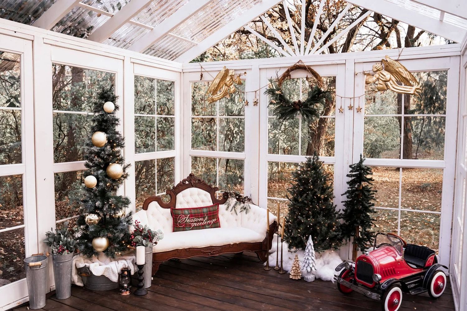 A festive sunroom decorated with Christmas trees, a red bench, wreath, and vintage toy car surrounded by large windows.