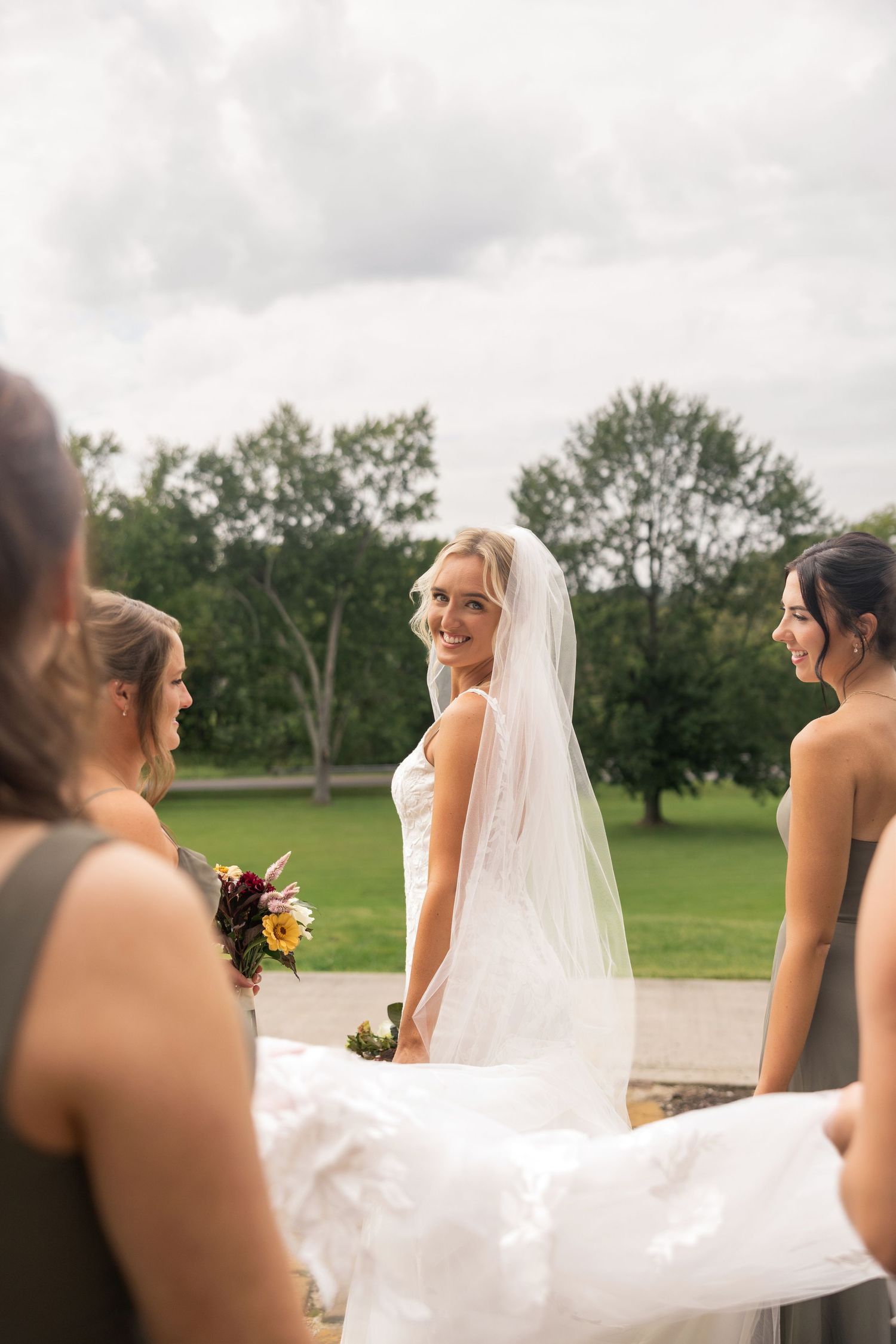 Smiling bride in white gown and veil with bridesmaids in olive dresses holding her train outdoors.